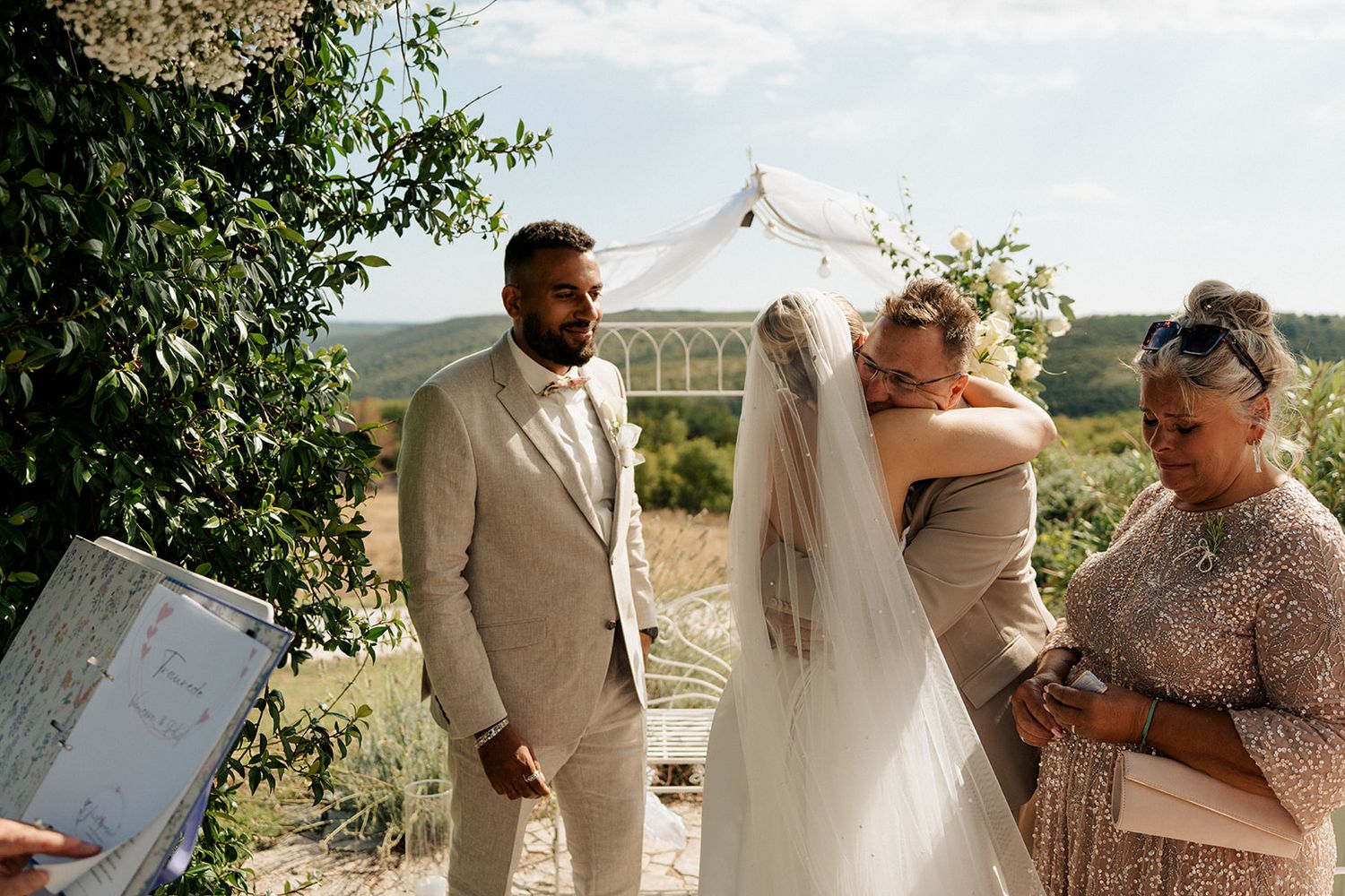 Bride and groom share an emotional hug during outdoor wedding ceremony with guests and scenic landscape in background.