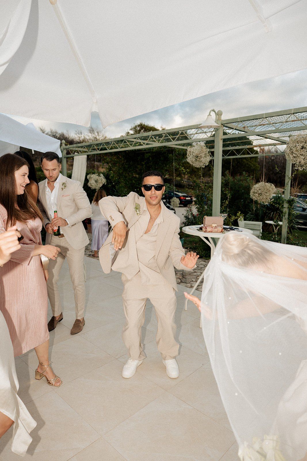 Groom in beige suit and sunglasses dances energetically at outdoor wedding reception under white tent.