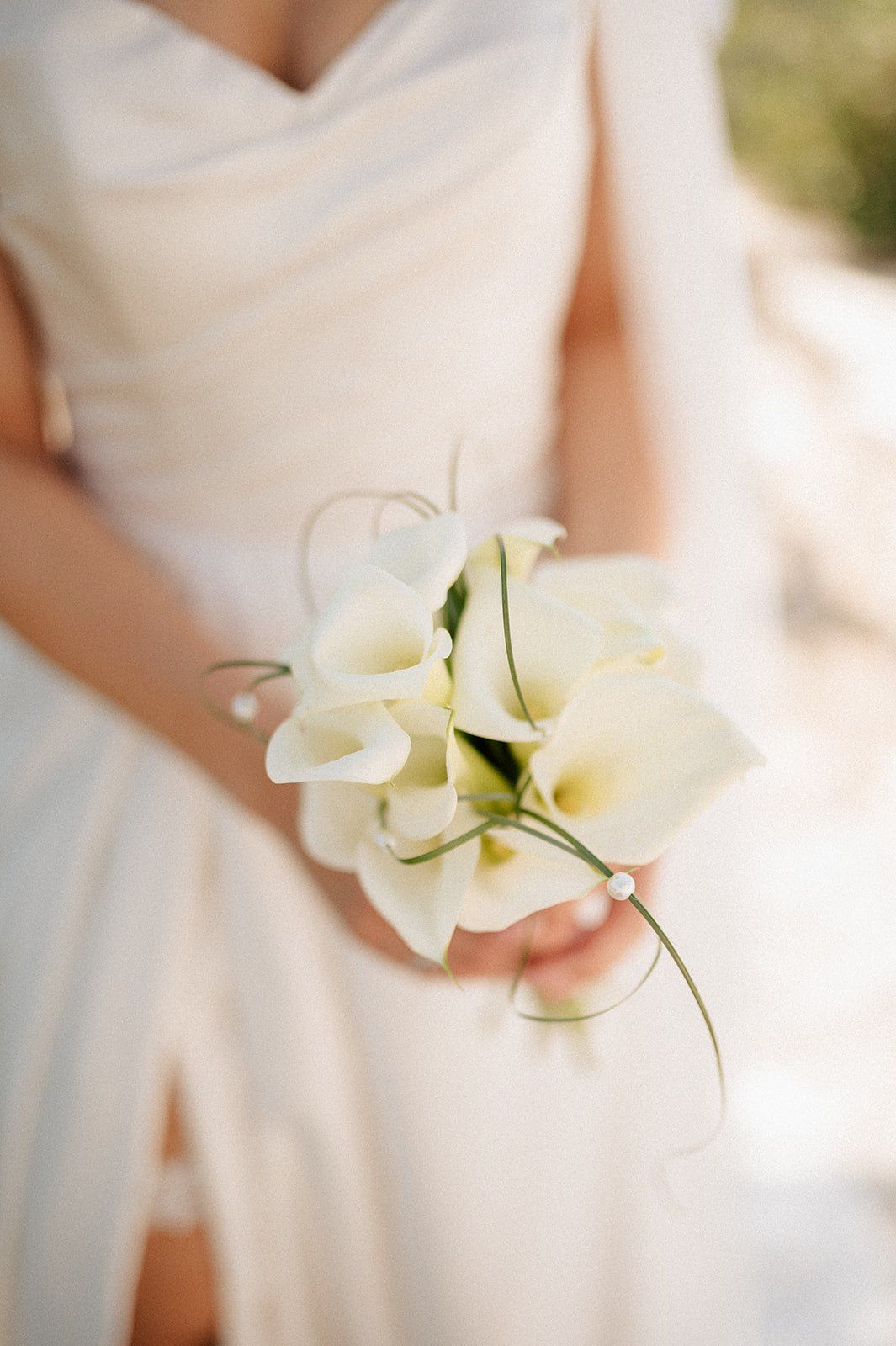 Bride in white off-shoulder wedding gown holding elegant white calla lily bouquet with green stems and decorative curls.