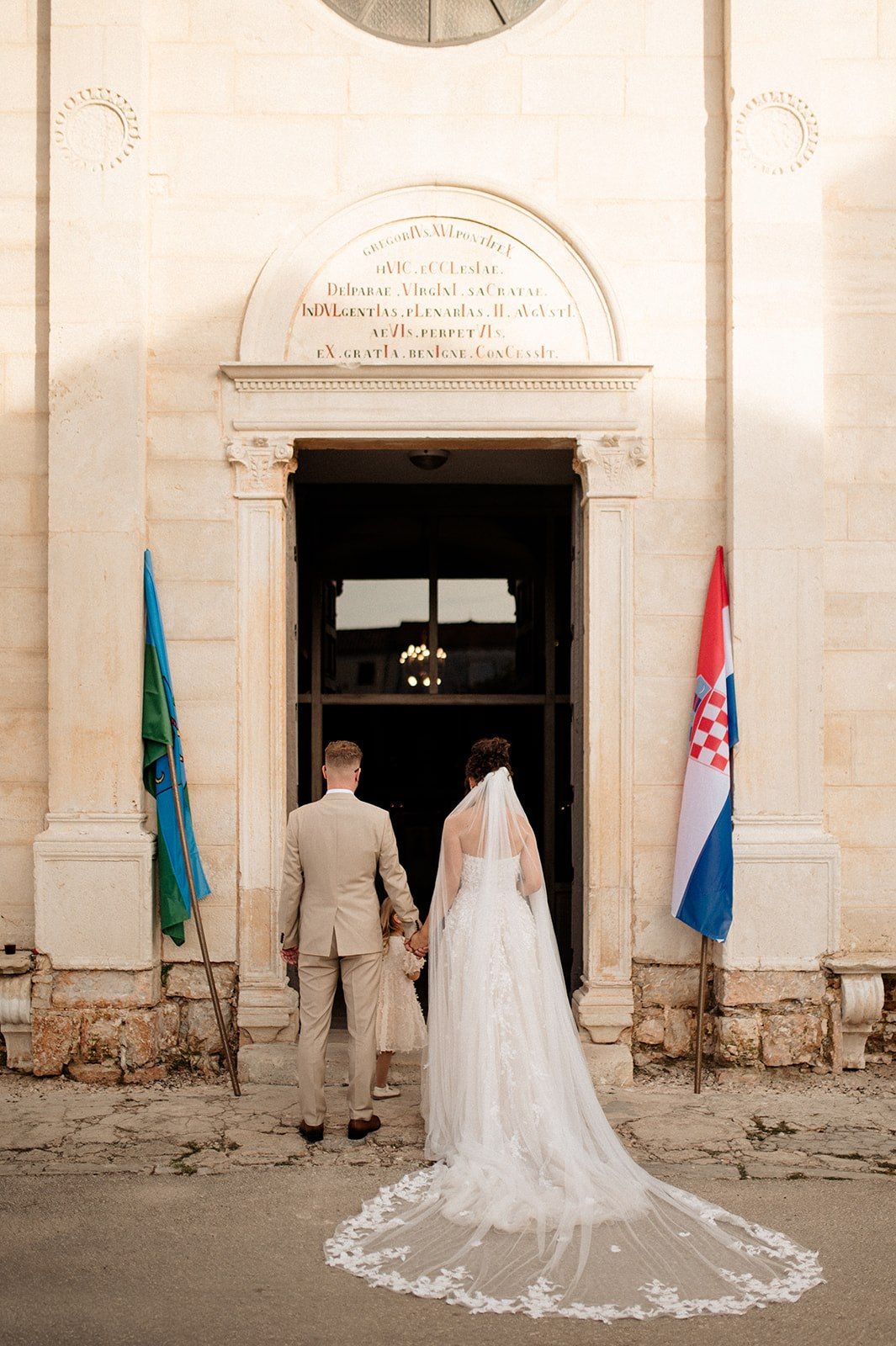 Bride and groom entering a historic stone church in Croatia, with a flowing lace veil and Croatian flag displayed outside.