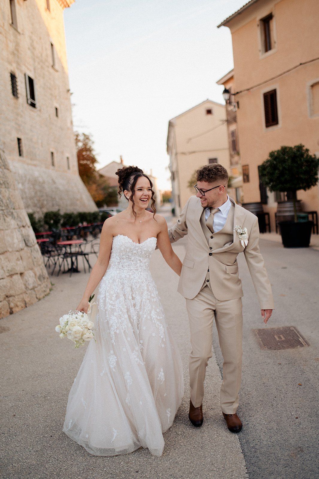 A joyful bride in a white lace gown and groom in a beige suit walk together through a European stone village street.