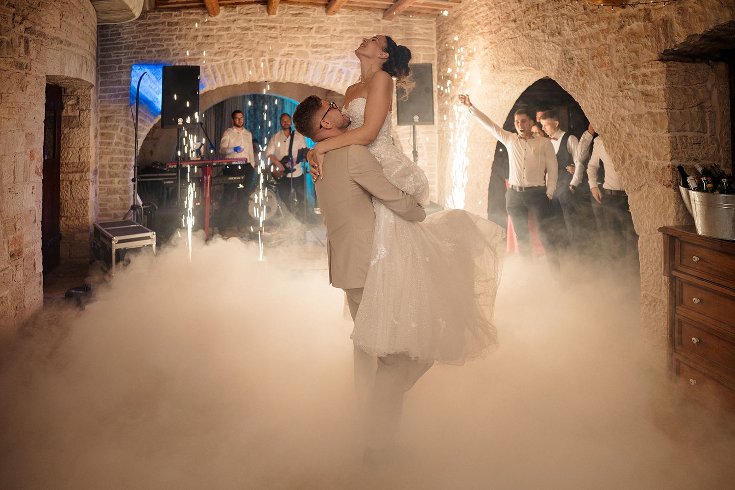 Bride and groom share first dance surrounded by dramatic fog and sparkling fountains in a rustic stone venue.