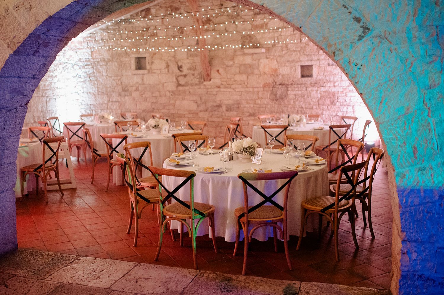 Elegant dining room in a rustic brick vaulted cellar with round tables, white linens, and colorful wooden chairs.