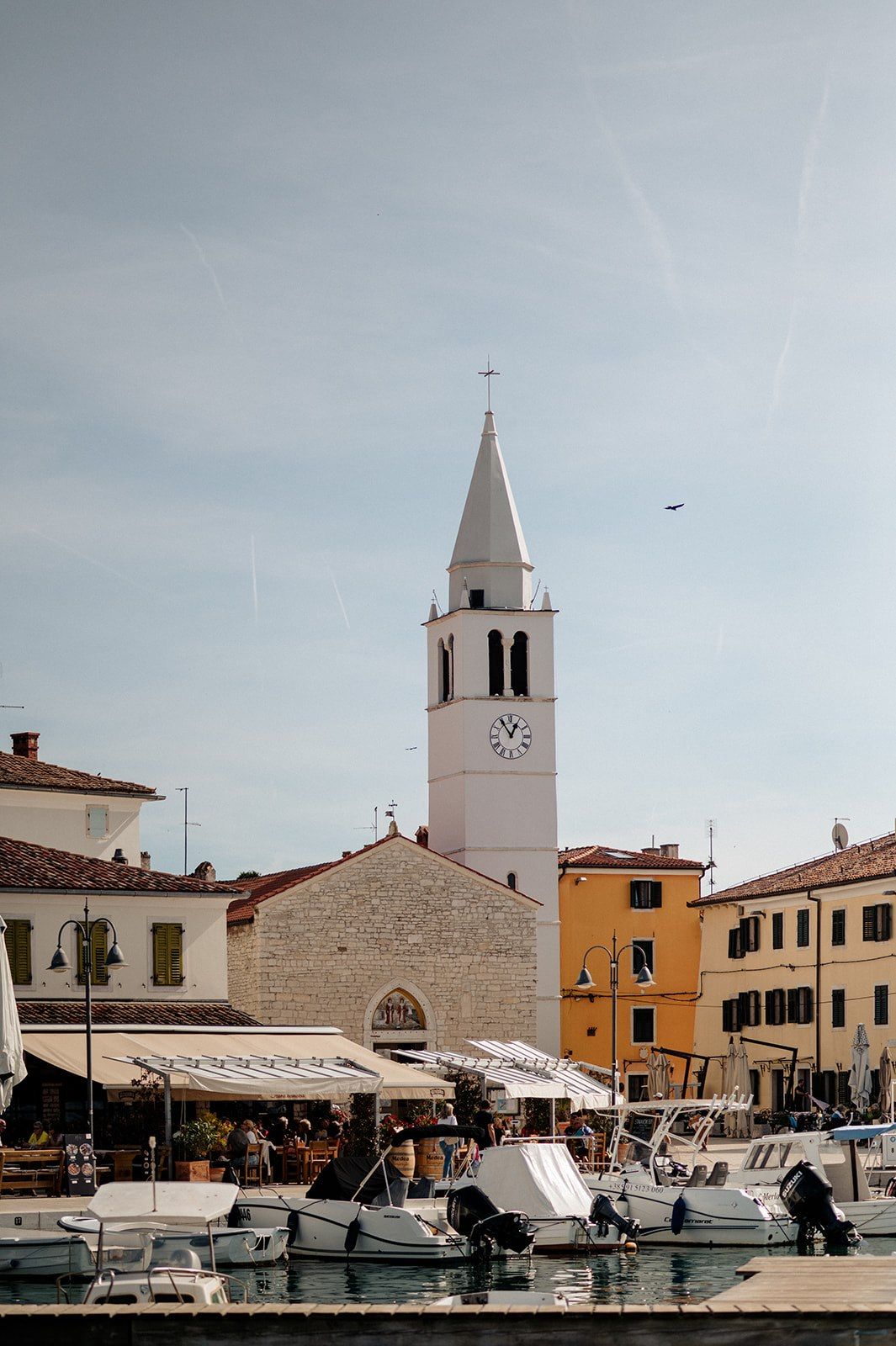 White church bell tower with clock face overlooks a small marina with boats in a Mediterranean coastal town.