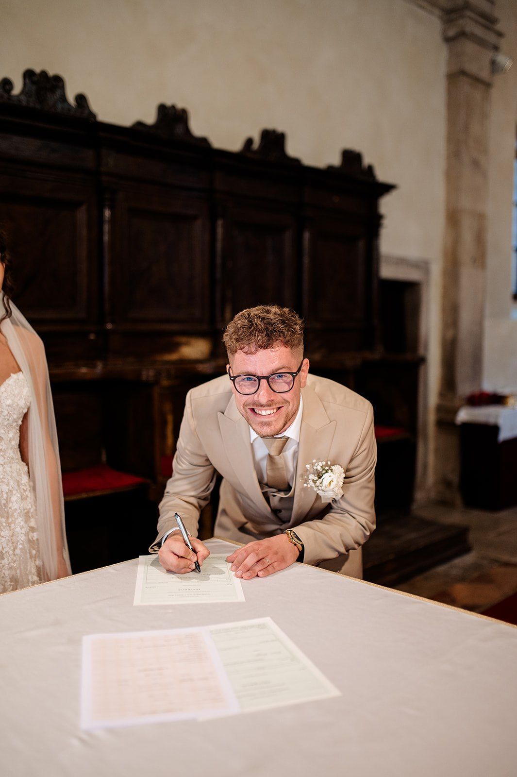 Smiling groom in tan suit signing wedding register at ceremony table.