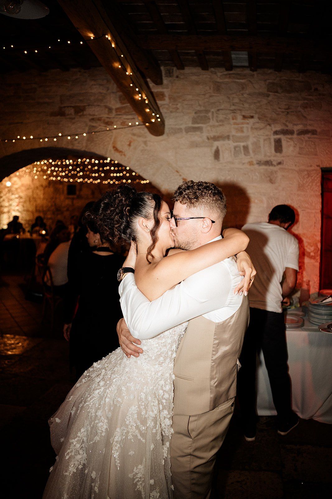 Newlyweds share a romantic first dance kiss in a warmly lit stone venue adorned with twinkling fairy lights.