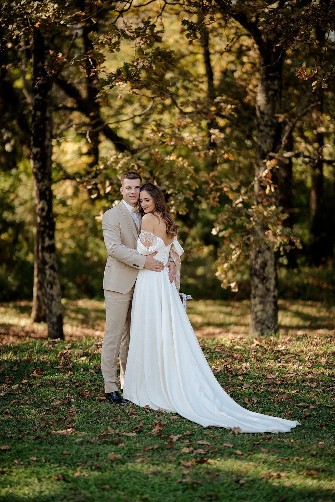 Bride and groom embracing in autumn forest, she in white gown, he in beige suit, surrounded by golden foliage.