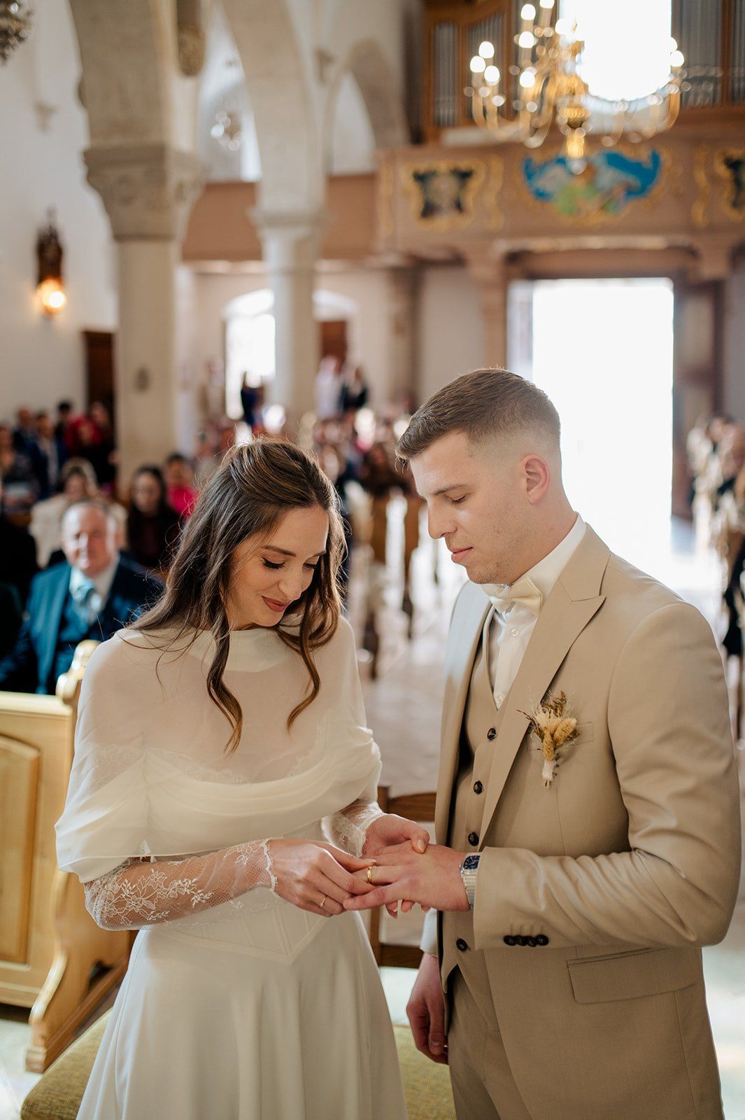 Groom placing wedding ring on bride's finger during church ceremony, both dressed elegantly in white and beige attire.