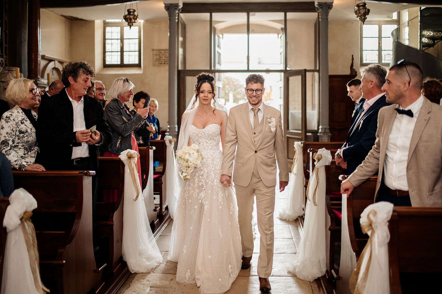 Bride and groom walk down church aisle together, surrounded by smiling guests seated in wooden pews.