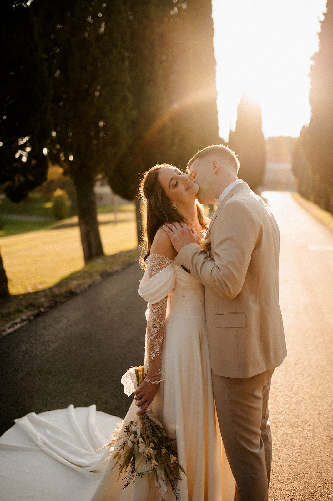 Bride and groom share a romantic kiss at golden hour on a tree-lined path, bride holding a dried floral bouquet.