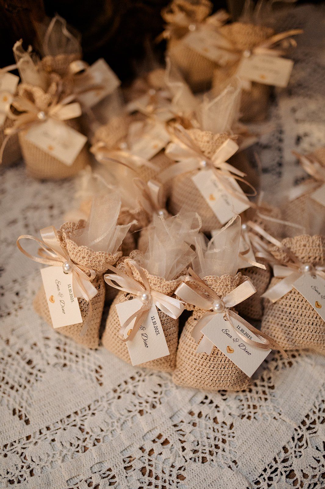 Rustic burlap wedding favor bags tied with ribbon and gift tags displayed on lace tablecloth.