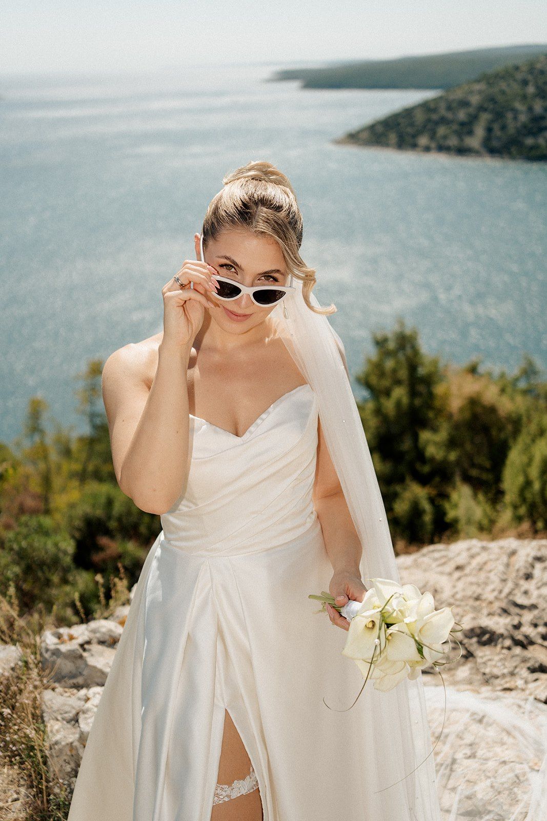 Bride in white wedding dress and sunglasses holding bouquet with scenic ocean and green hills backdrop.