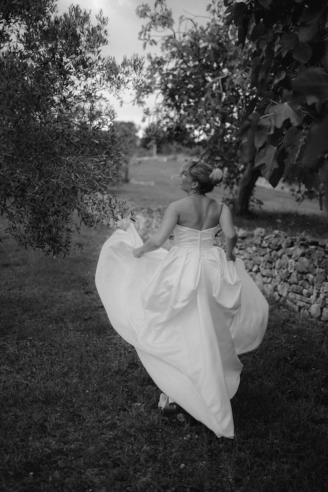 Black and white photo of bride in ball gown walking through garden, dress flowing dramatically.