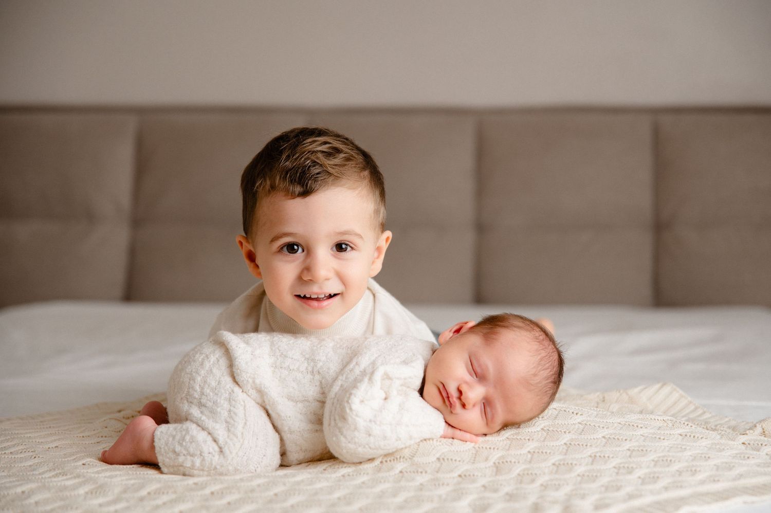 Two siblings on a textured bed, one wearing a white sweater sitting up while the newborn baby sleeps beside them.