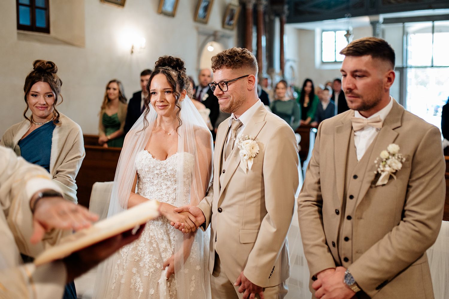 Bride and groom holding hands at altar during wedding ceremony in church, with guests and best man standing nearby.