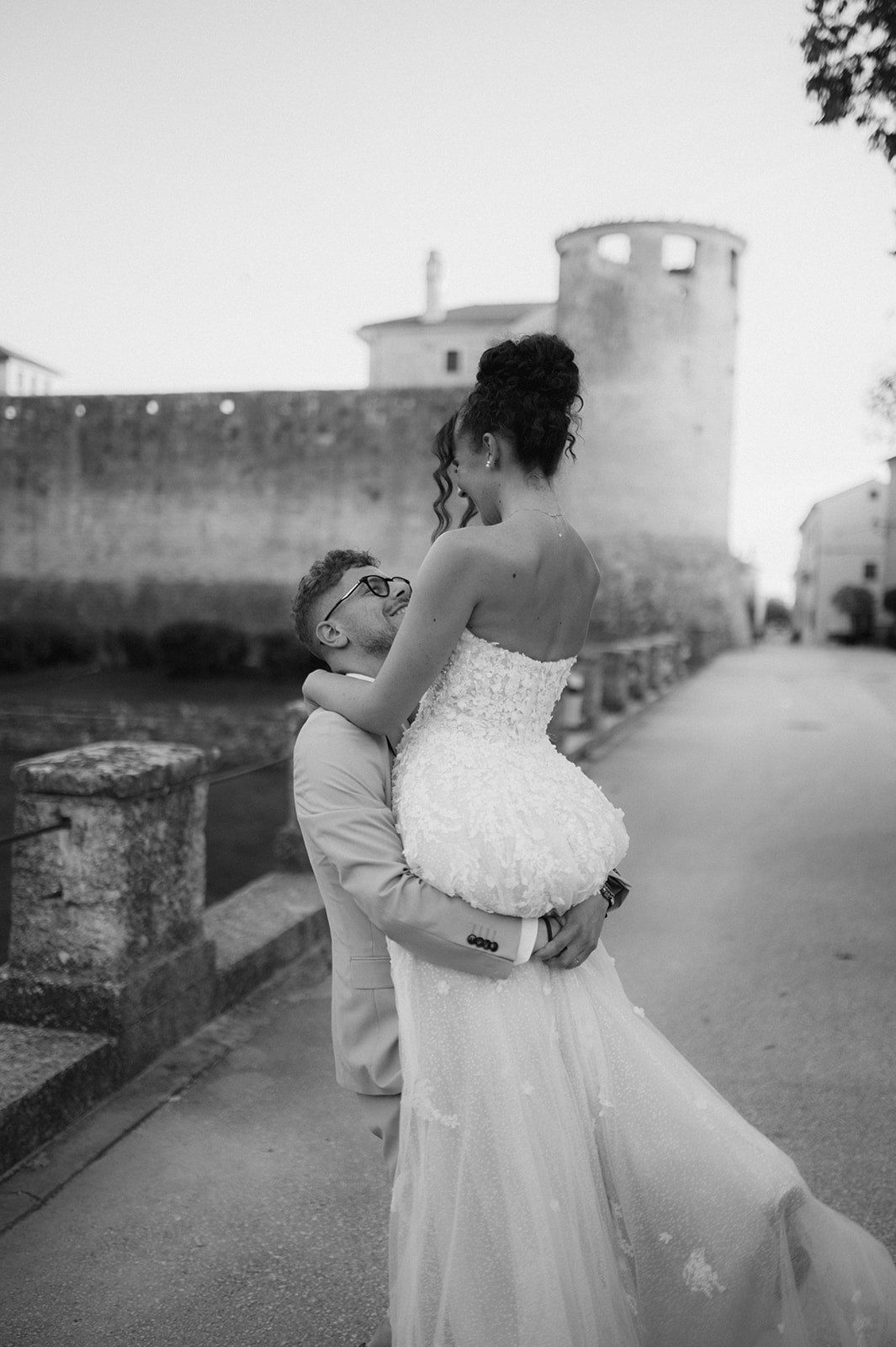 Black and white photo of a groom lifting his bride in a white gown near a historic stone wall.