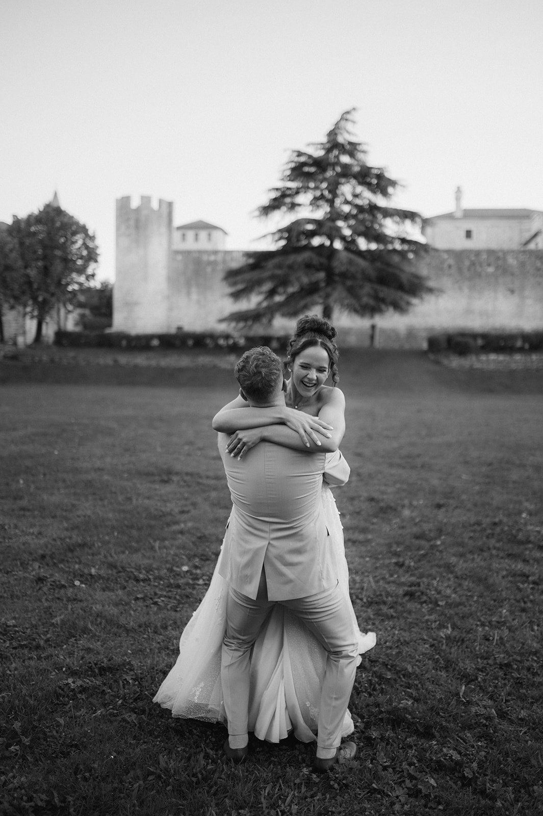 Black and white photo of bride and groom embracing joyfully in front of a castle.