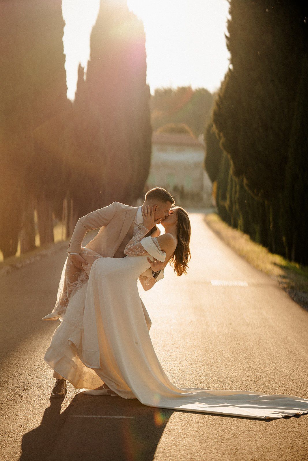 Bride and groom sharing a romantic dip kiss on a sunlit tree-lined road during golden hour wedding photography.