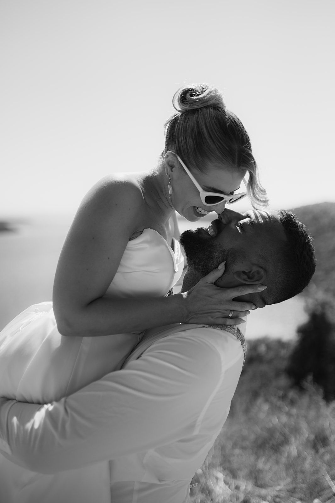 Black and white photo of a romantic couple sharing a kiss outdoors, woman in white dress with sunglasses, scenic coastal backdrop.