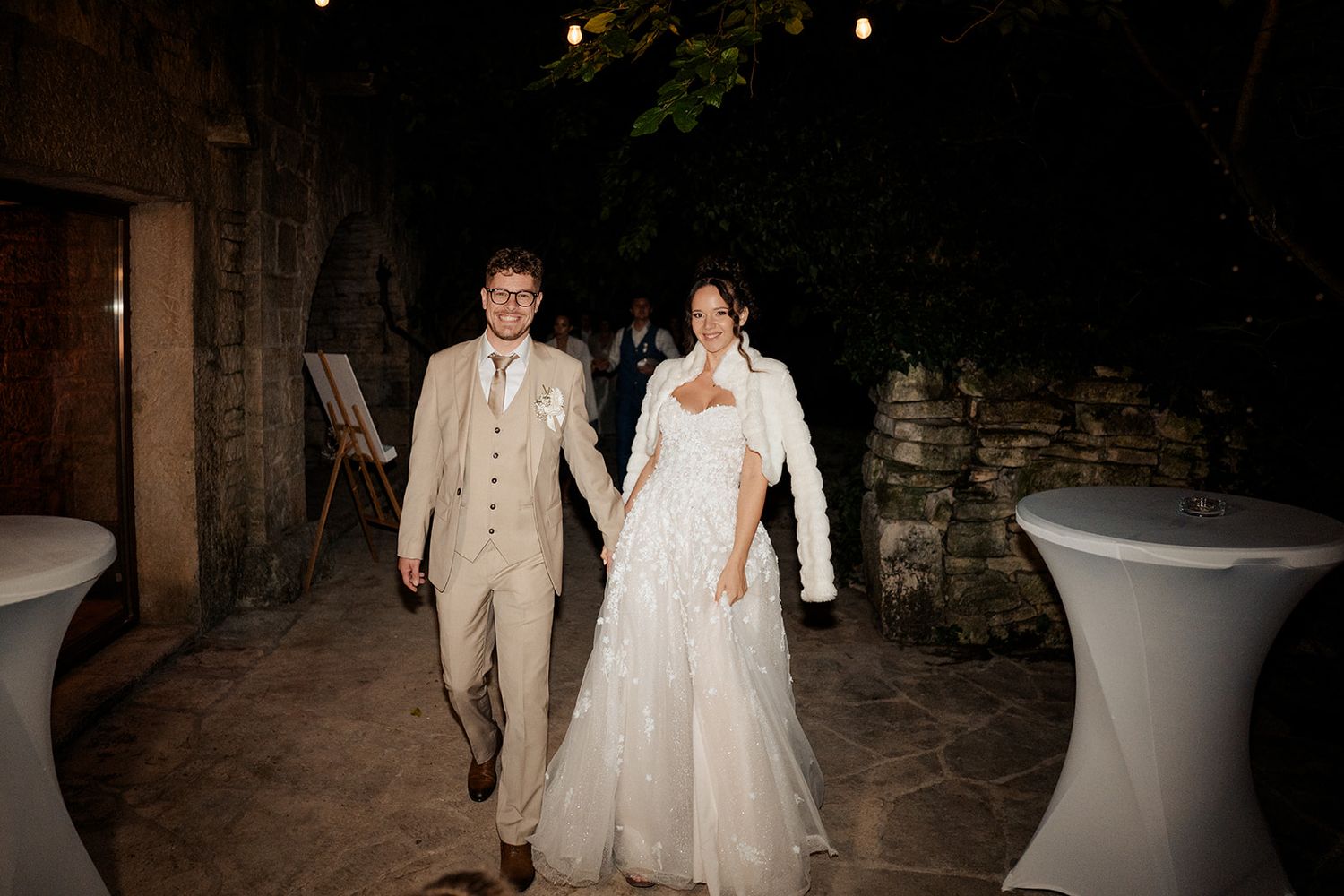 Bride and groom walk hand in hand at an outdoor evening wedding reception with string lights and stone walls.