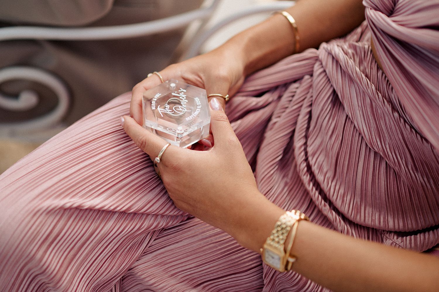 Woman in pleated mauve dress holding a perfume bottle, wearing elegant gold jewelry and bracelets.