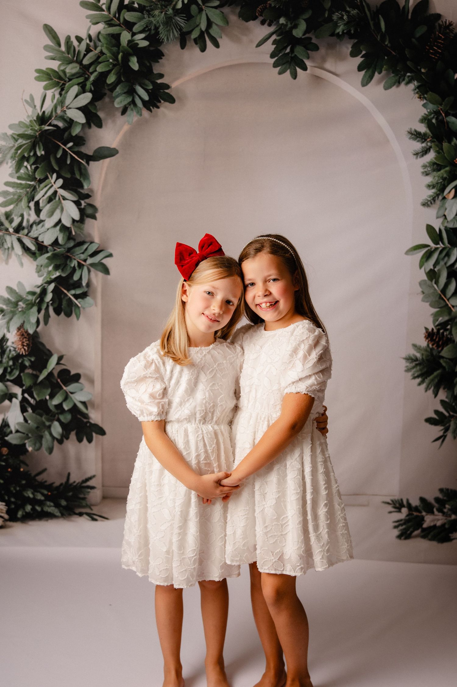 Two children wearing white lace dresses pose together against a white backdrop with green foliage decorations.