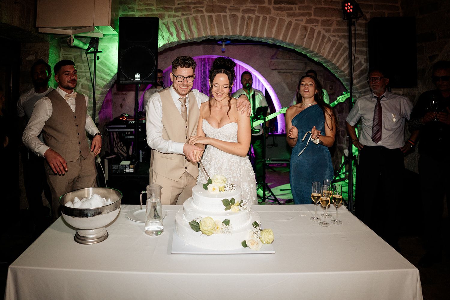 Bride and groom cutting wedding cake together at elegant reception venue with guests and band in background.