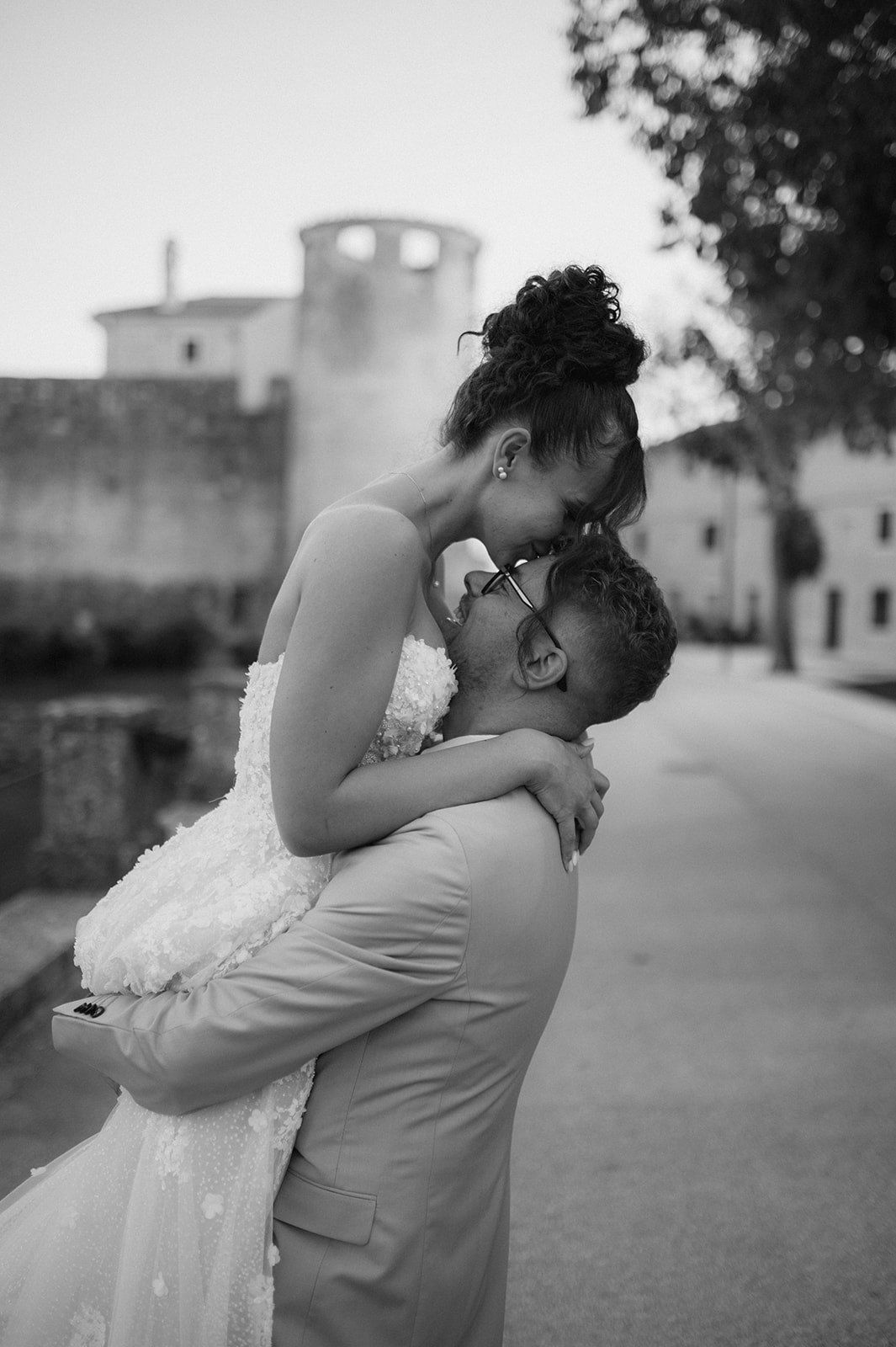 Black and white photo of a bride and groom embracing outdoors, sharing a tender moment near a historic stone building.
