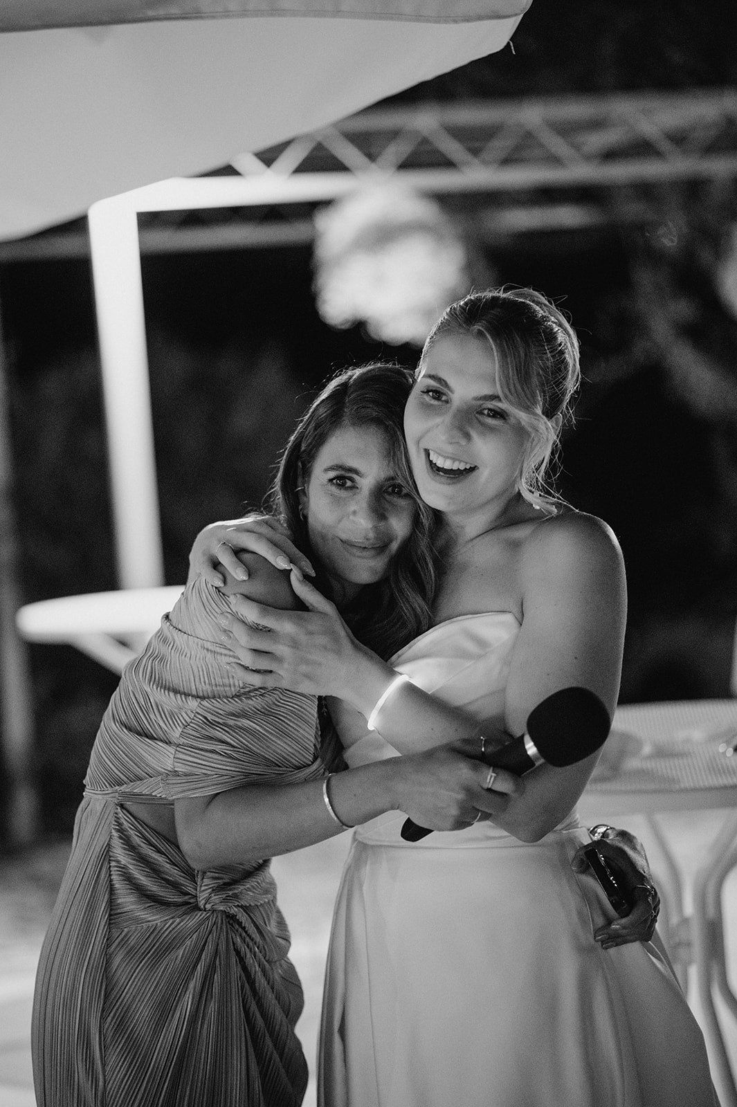 Black and white photo of smiling bride in strapless gown hugging guest while holding microphone at reception.