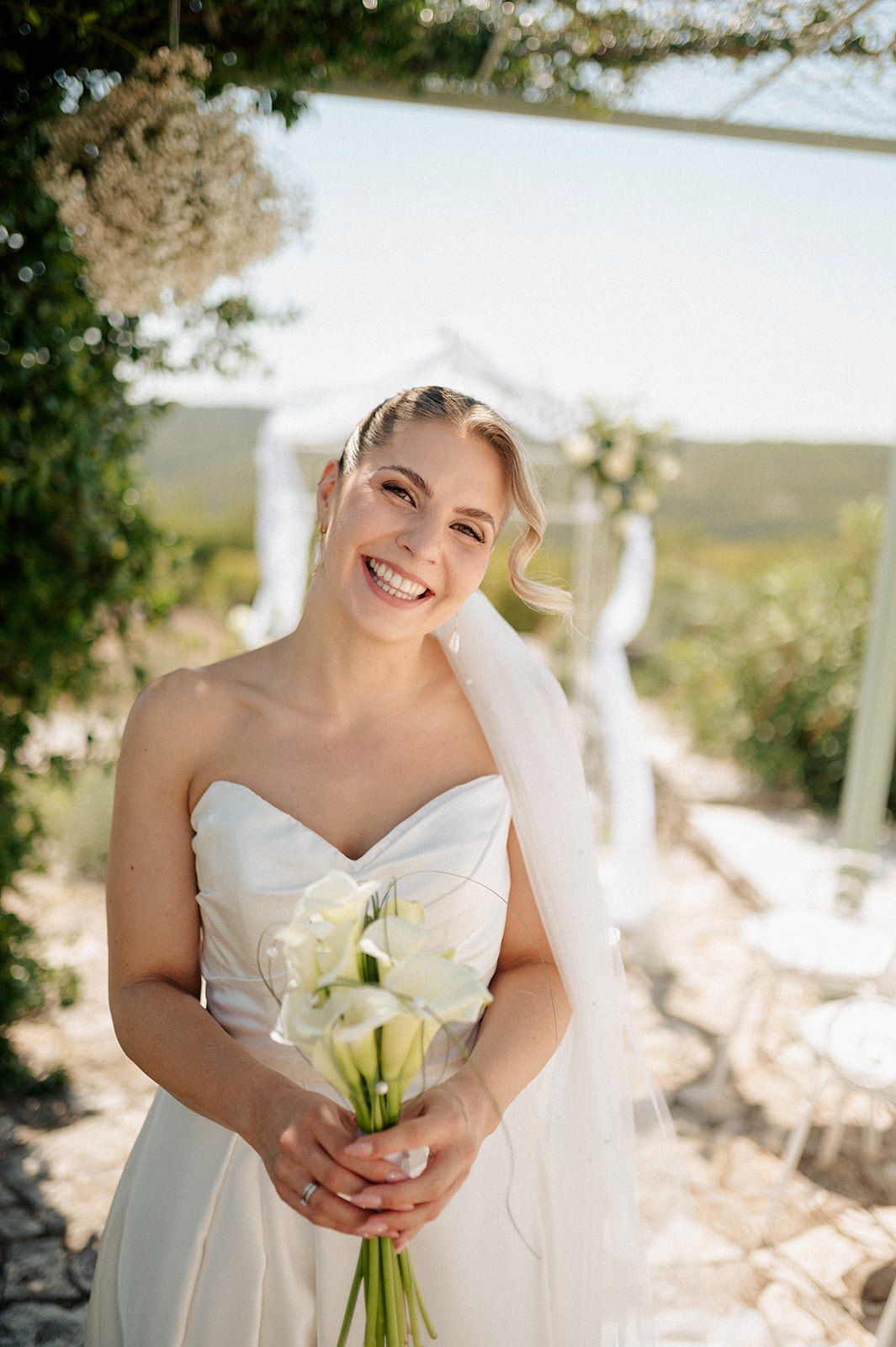 Smiling bride in strapless white wedding dress holding white calla lily bouquet outdoors with veil and vineyard background.