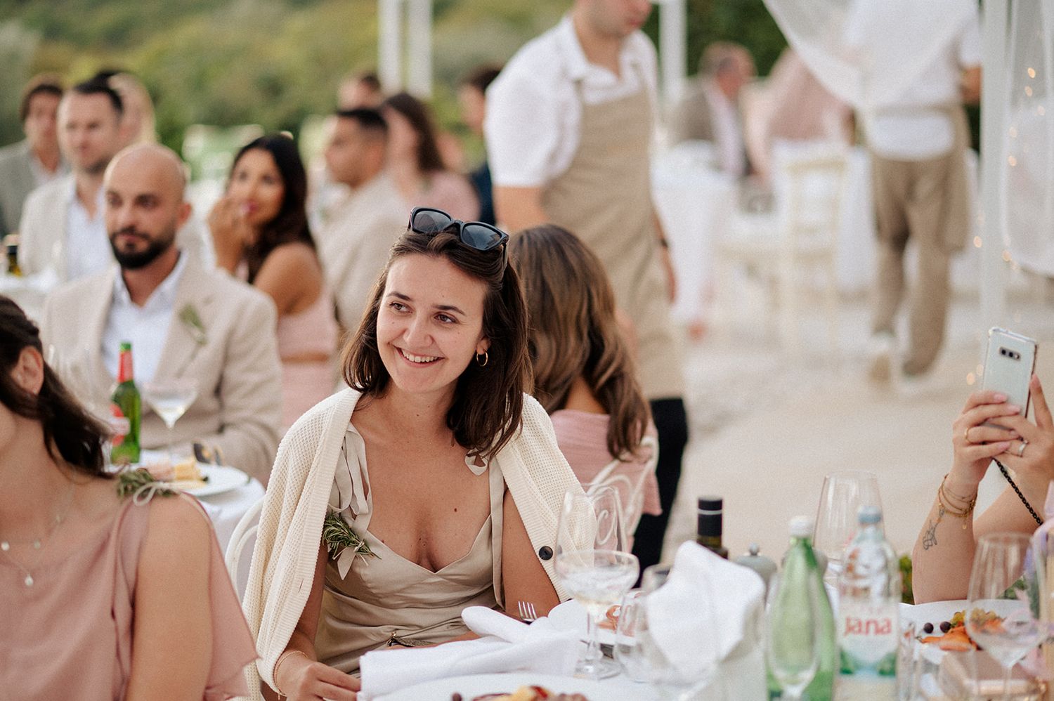 Woman smiling at an outdoor wedding reception dinner, seated at a decorated table with guests in the background.