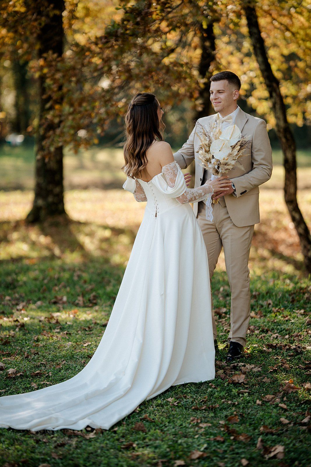Bride in white off-shoulder gown and groom in beige suit share a romantic moment in an autumn park.