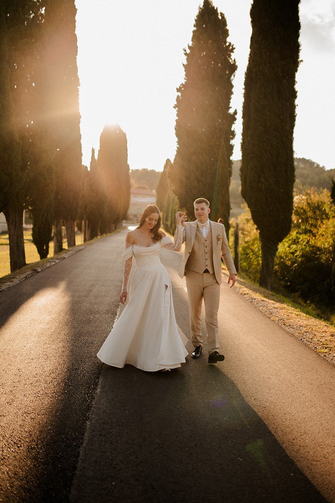 Bride and groom walking down a sunlit cypress-lined road in Tuscany during golden hour.
