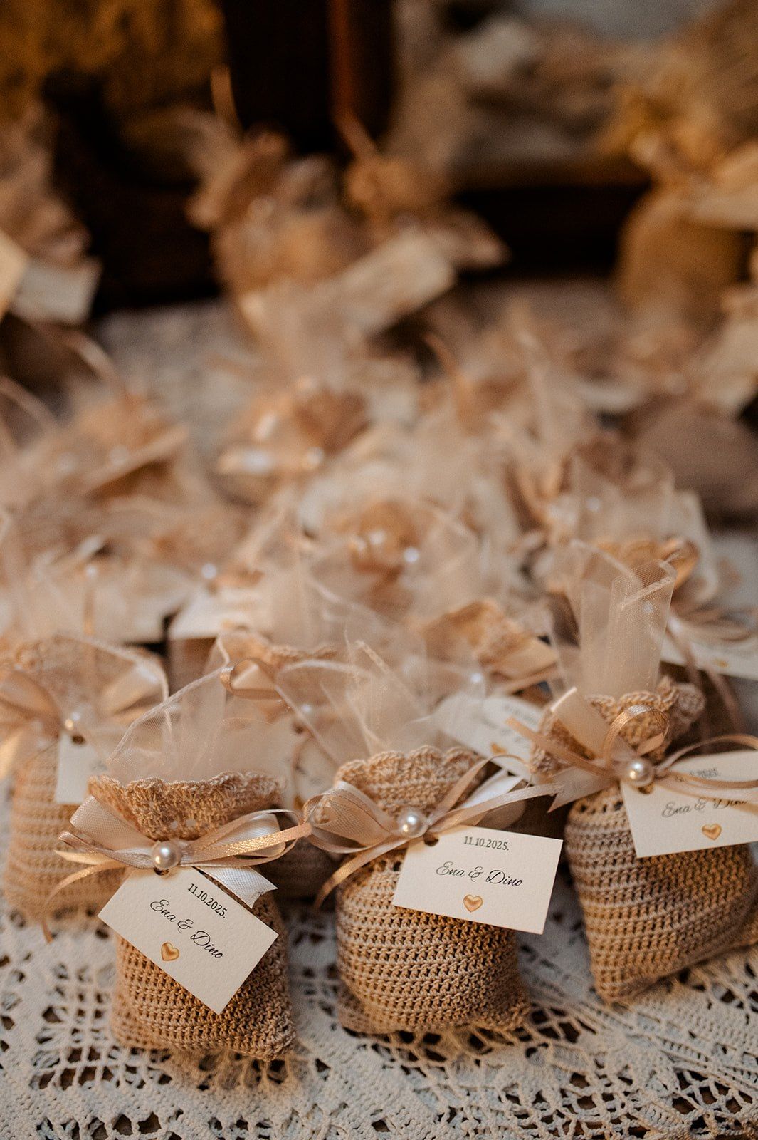 Rustic burlap wedding favor bags tied with sheer ribbon and personalized tags displayed on lace tablecloth.