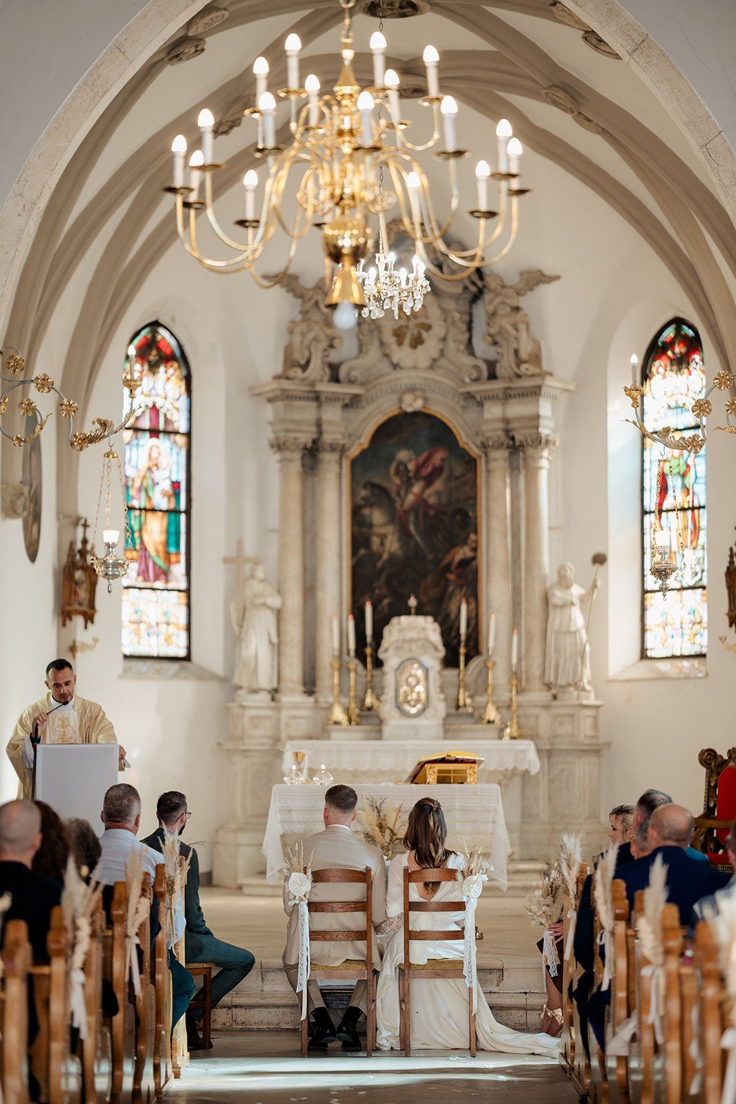 Elegant church wedding ceremony with couple at baroque altar, stained glass windows, and golden chandelier overhead.