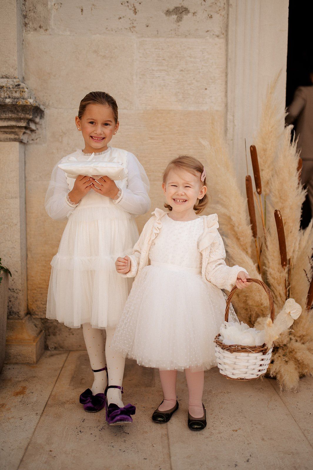 Two young flower girls in white tulle dresses pose outdoors, one holding a pillow and the other a wicker basket.