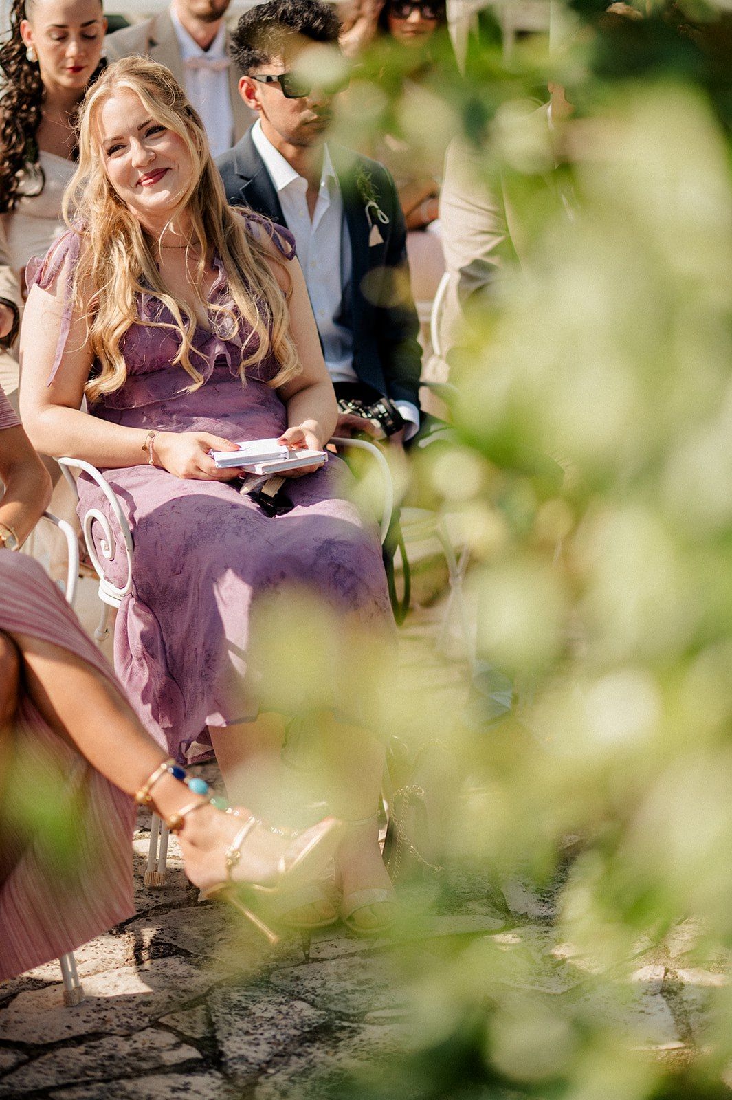 Smiling wedding guest in purple dress seated outdoors, photographed through green foliage.