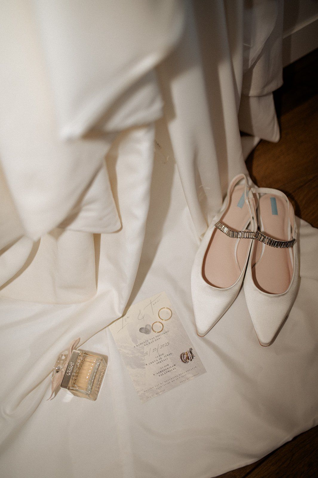 White bridal flat shoes with crystal strap beside wedding invitation, gold rings, and garter on a white dress.
