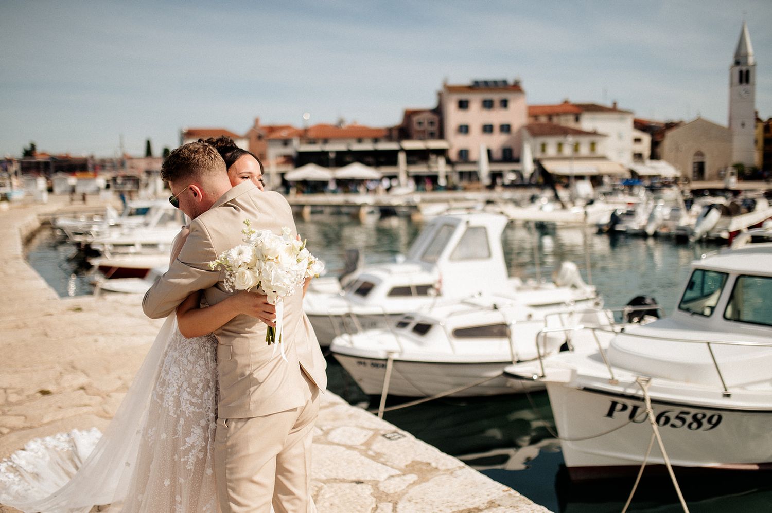 Bride and groom embrace on a sunny marina waterfront in Croatia, with white boats and historic buildings behind them.