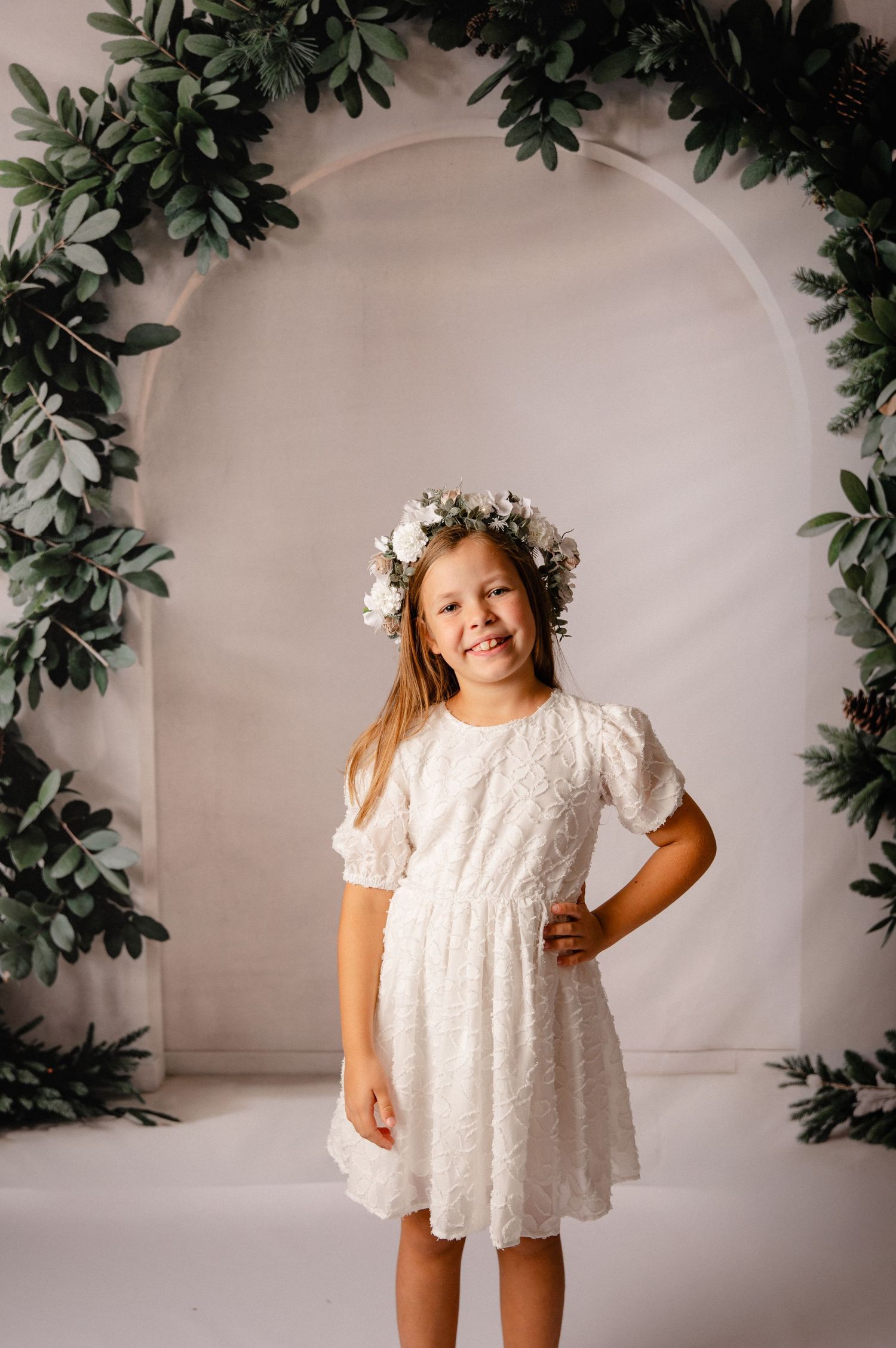 A young child in a white lace dress and floral crown poses against a backdrop with green foliage decorations.