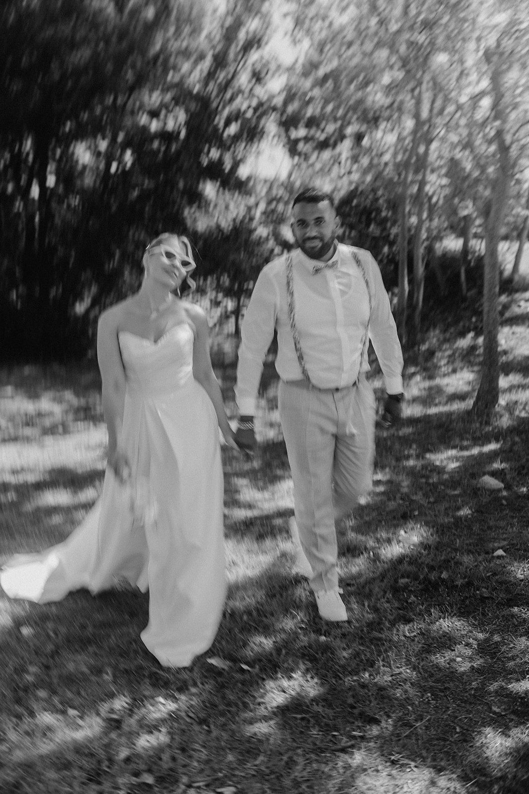 Bride and groom holding hands while walking through a sunlit forest in black and white wedding photography.