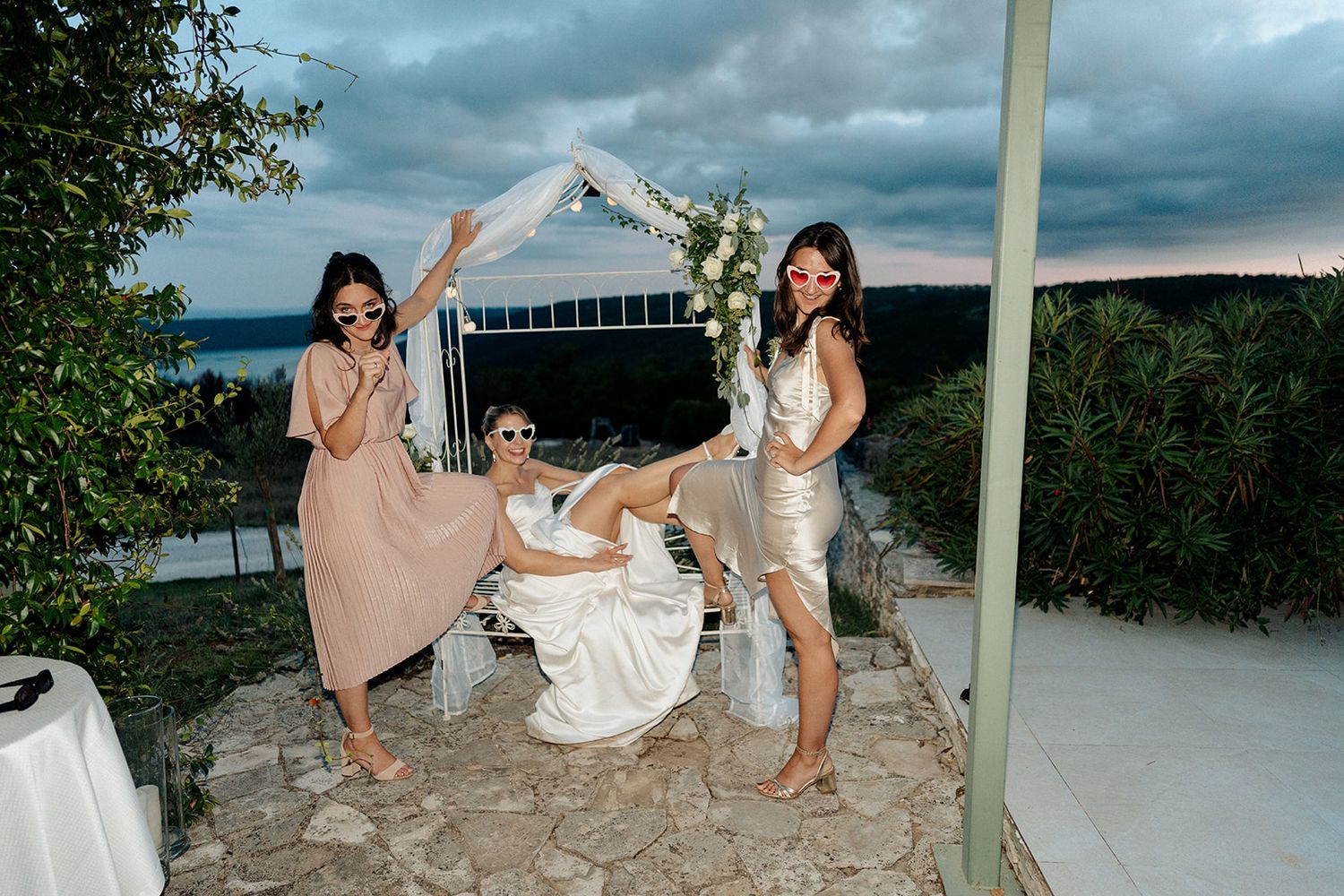 Bride and two bridesmaids posing playfully under floral arch at outdoor evening wedding celebration.
