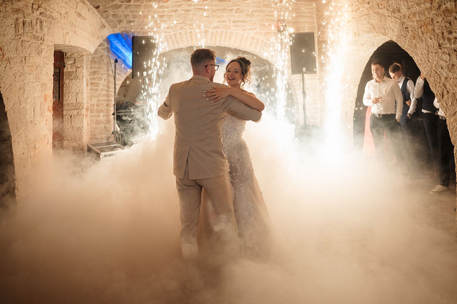 Bride and groom share first dance on fog-covered floor in rustic stone venue with sparkling lights.
