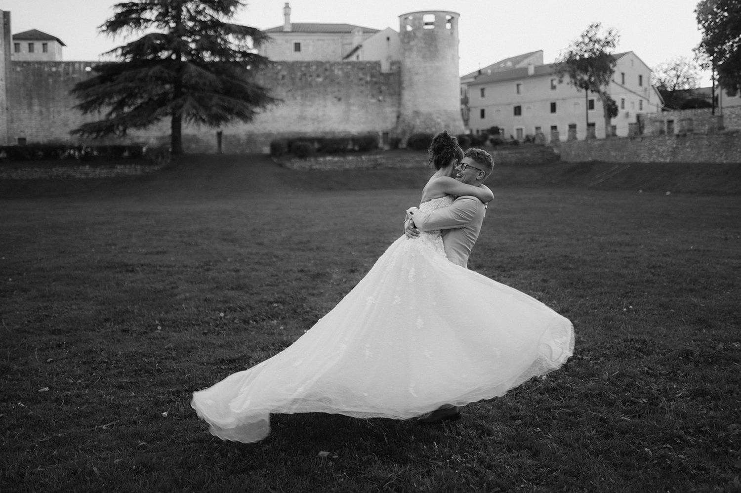Black and white photo of bride and groom embracing in field near historic stone castle walls.