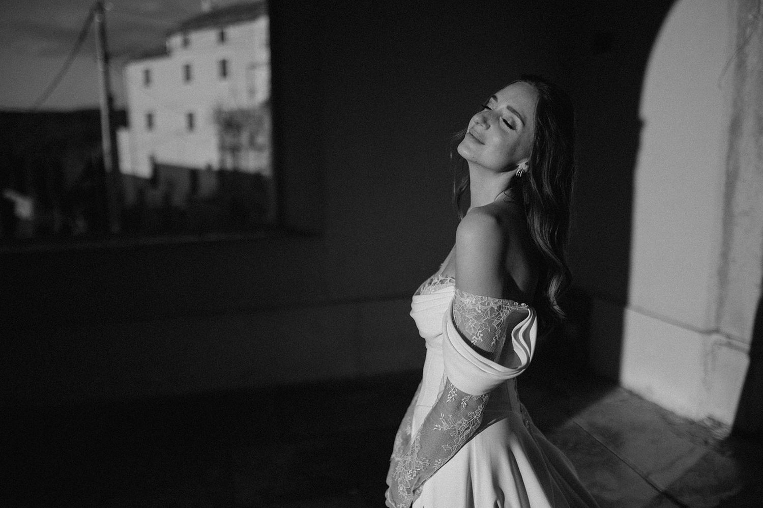 Black and white photo of a woman in an off-shoulder white gown posing gracefully outdoors near a historic building.