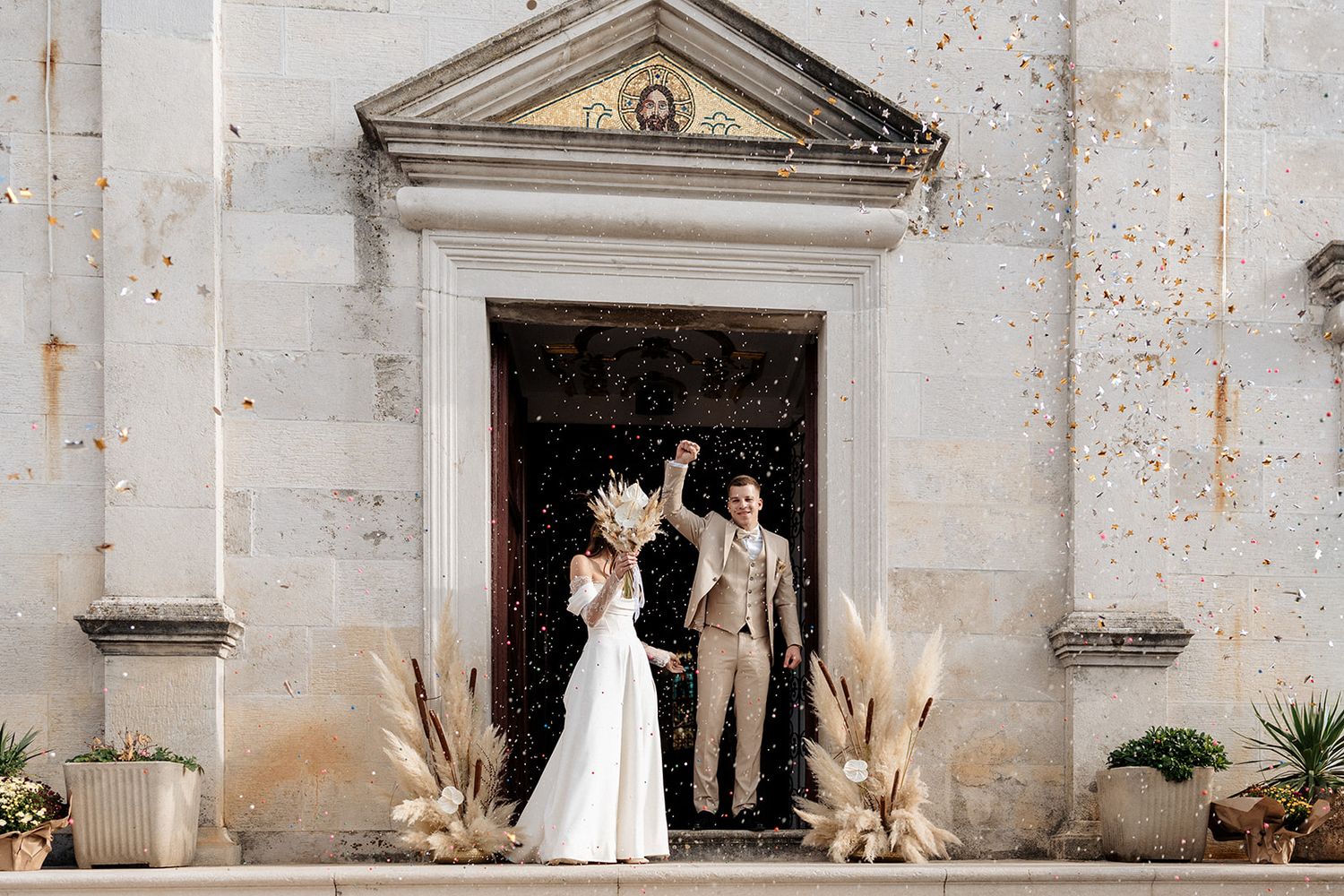 Newlywed couple celebrating outside a rustic stone church entrance with pampas grass decor and confetti.