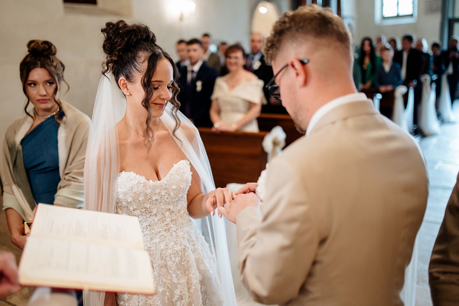 Bride and groom exchange rings at church altar during wedding ceremony, with guests watching in background.