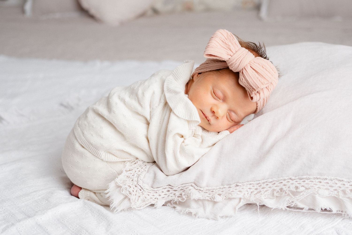 Newborn baby sleeping peacefully on soft white bedding wearing delicate lace clothing.