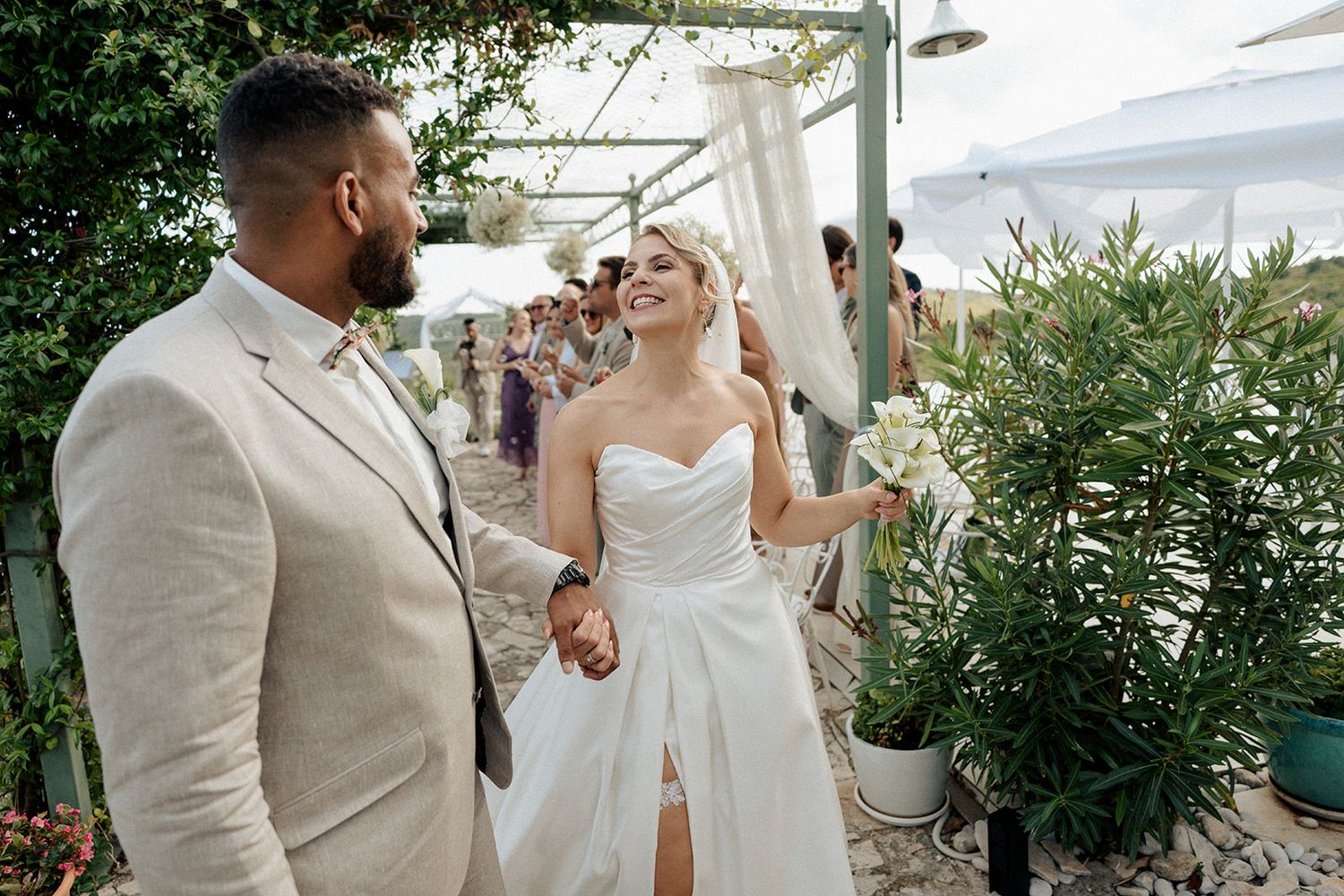 Smiling bride in white strapless gown holds hands with groom in beige suit at outdoor wedding ceremony.
