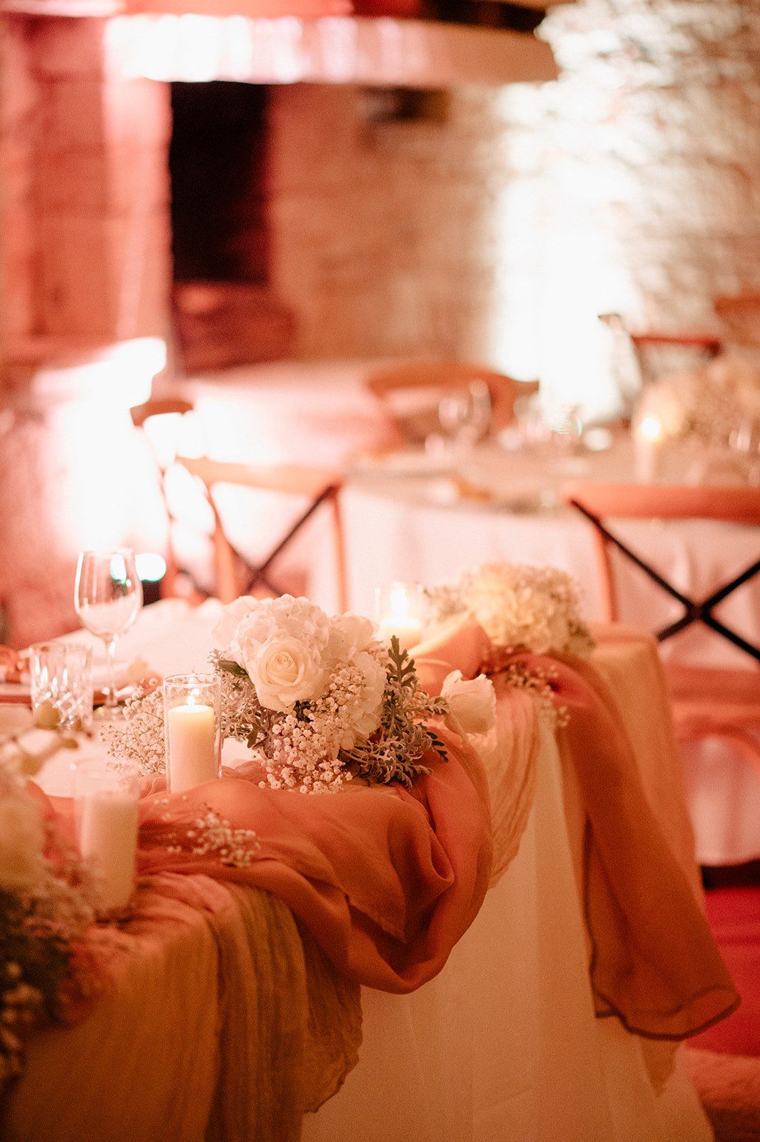 Elegant wedding reception table with blush draping, white roses, candles, and warm romantic lighting in a rustic venue.